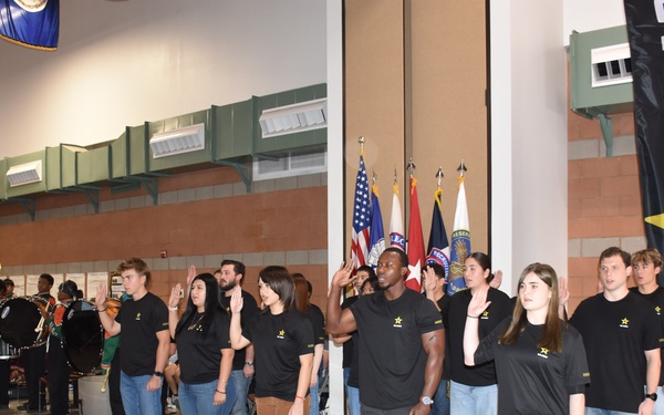 A U.S. Army enlistment ceremony is taking place in what appears to be a large indoor facility such as an armory or gymnasium. A uniformed soldier in Army Combat Uniform (ACU) is administering the oath of enlistment to a group of approximately 12-15 recruits, all wearing matching black U.S. Army t-shirts with the Army star logo. A military band and color guard with multiple flags are visible in the background.