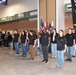A U.S. Army enlistment ceremony is taking place in what appears to be a large indoor facility such as an armory or gymnasium. A uniformed soldier in Army Combat Uniform (ACU) is administering the oath of enlistment to a group of approximately 12-15 recruits, all wearing matching black U.S. Army t-shirts with the Army star logo. A military band and color guard with multiple flags are visible in the background.
