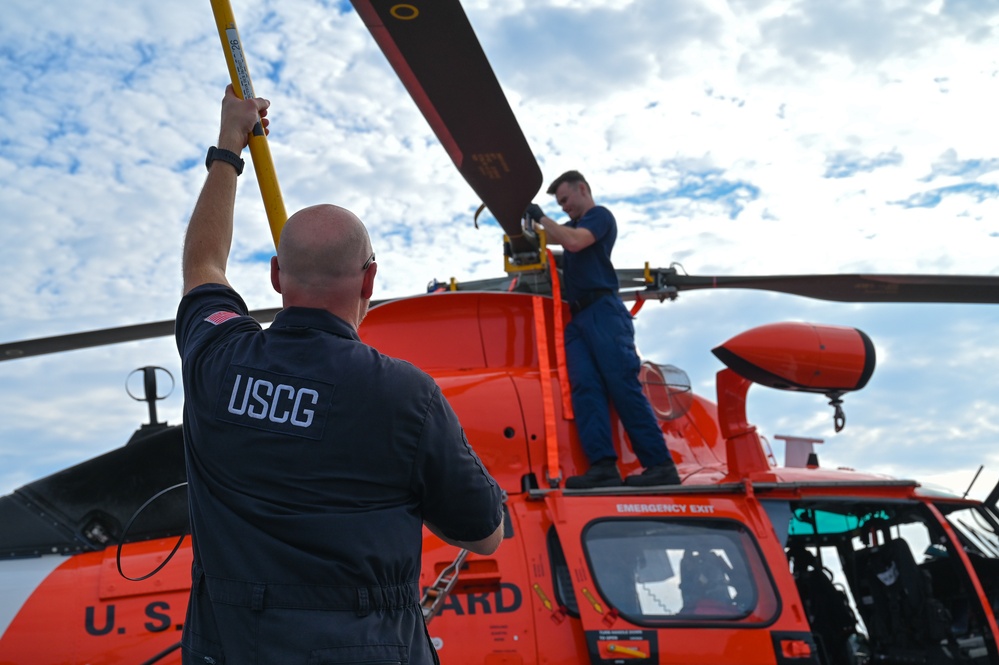 Two United States Coast Guard (USCG) personnel are performing maintenance or pre-flight checks on an orange Coast Guard helicopter. One crew member stands on top of the aircraft working on the rotor assembly, while another holds a yellow pole or tool upward toward the rotor head. The helicopter is clearly marked with 'U.S. COAST GUARD' and features the distinctive international orange color scheme.