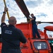 Two United States Coast Guard (USCG) personnel are performing maintenance or pre-flight checks on an orange Coast Guard helicopter. One crew member stands on top of the aircraft working on the rotor assembly, while another holds a yellow pole or tool upward toward the rotor head. The helicopter is clearly marked with 'U.S. COAST GUARD' and features the distinctive international orange color scheme.
