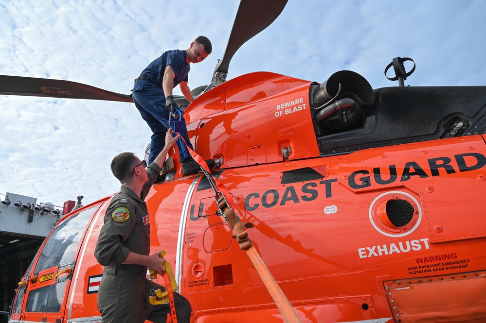 Two personnel are working on a bright orange U.S. Coast Guard helicopter, likely securing or maintaining it on a ship's deck. One person is standing on top of the aircraft while the other stands alongside, and they appear to be handling orange tie-down straps. The helicopter displays multiple safety markings including 'BEWARE OF BLAST,' 'EXHAUST,' and 'WARNING HIGH PRESSURE EMERGENCY FLOTATION GEAR STOWAGE.'