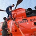 Two personnel are working on a bright orange U.S. Coast Guard helicopter, likely securing or maintaining it on a ship's deck. One person is standing on top of the aircraft while the other stands alongside, and they appear to be handling orange tie-down straps. The helicopter displays multiple safety markings including 'BEWARE OF BLAST,' 'EXHAUST,' and 'WARNING HIGH PRESSURE EMERGENCY FLOTATION GEAR STOWAGE.'