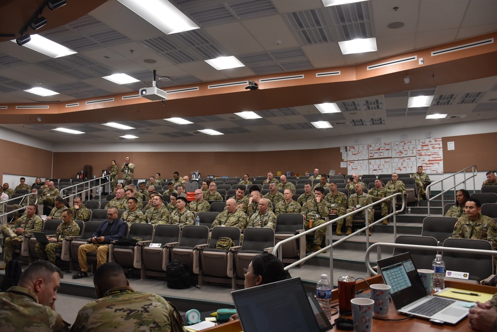 A large military briefing or training session is taking place in a tiered auditorium-style classroom. The majority of attendees are wearing U.S. Army combat uniforms (ACU/OCP pattern), with a small number of civilians present. The setting appears to be a formal military education or command briefing facility.