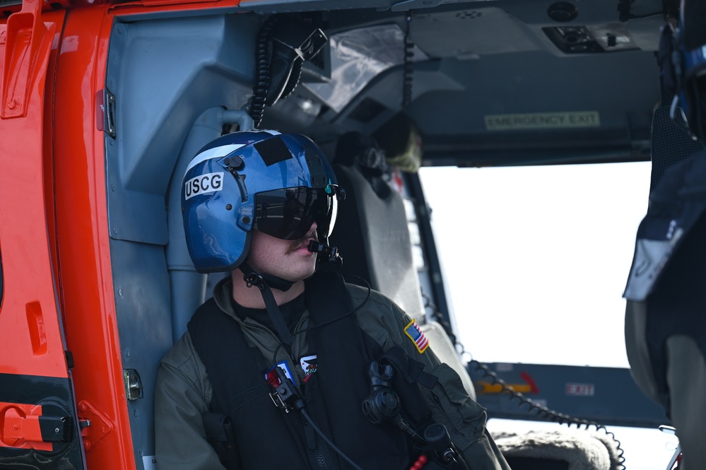 A United States Coast Guard (USCG) crew member is seated inside a helicopter, wearing a blue flight helmet marked 'USCG' and a flight suit with a US flag patch. The individual appears to be on an active mission or patrol, looking outward from the open helicopter door. The aircraft interior shows orange and gray panels with visible 'EMERGENCY EXIT' labeling.