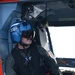 A United States Coast Guard (USCG) crew member is seated inside a helicopter, wearing a blue flight helmet marked 'USCG' and a flight suit with a US flag patch. The individual appears to be on an active mission or patrol, looking outward from the open helicopter door. The aircraft interior shows orange and gray panels with visible 'EMERGENCY EXIT' labeling.