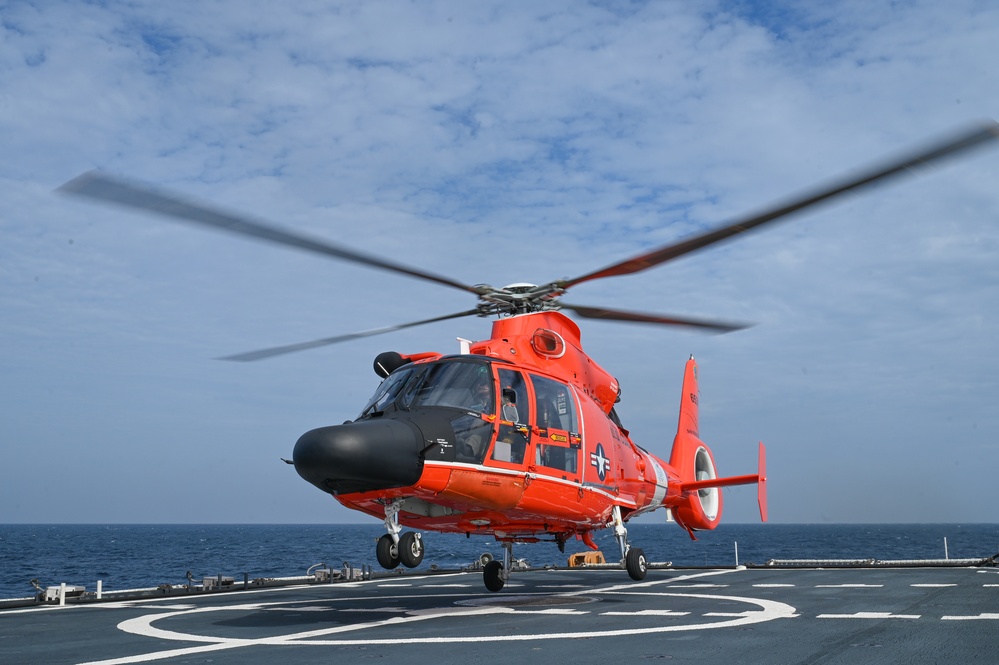 A bright orange U.S. Coast Guard helicopter, identifiable by its distinctive paint scheme and military star insignia, is landing or taking off from a ship's flight deck at sea. The rotor blades are in motion, visible as a blur above the aircraft. The setting is an open ocean environment under a partly cloudy sky.