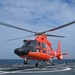 A bright orange U.S. Coast Guard helicopter, identifiable by its distinctive paint scheme and military star insignia, is landing or taking off from a ship's flight deck at sea. The rotor blades are in motion, visible as a blur above the aircraft. The setting is an open ocean environment under a partly cloudy sky.