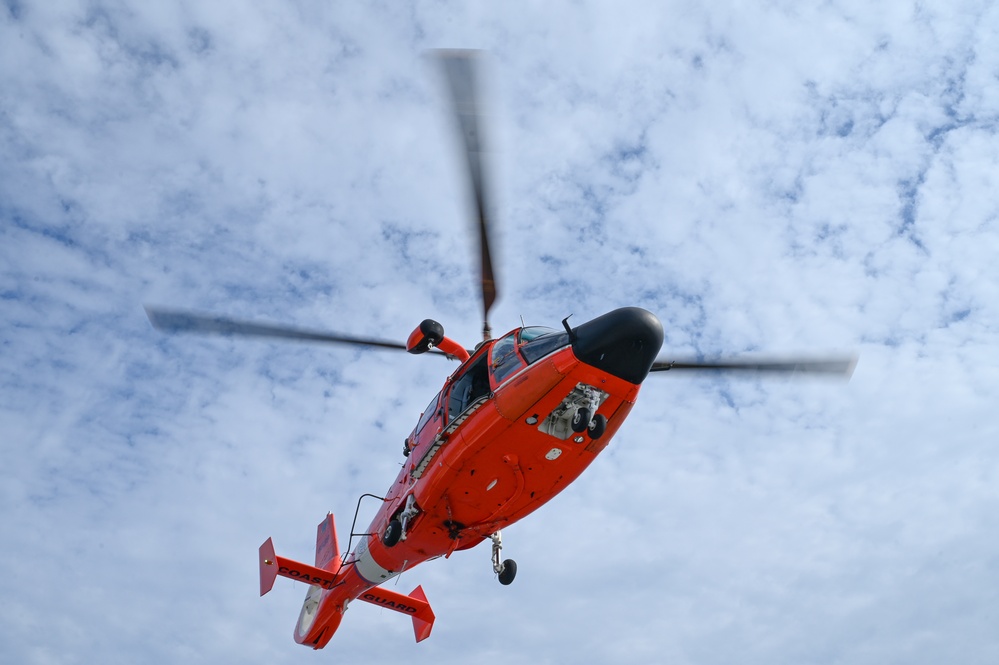 A bright orange Coast Guard helicopter is photographed from below against a partly cloudy blue sky. The image captures the aircraft in flight with its main rotor blades in motion, creating a blur effect. The text 'COAST GUARD' is visible on the underside of the tail section.