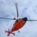 A bright orange Coast Guard helicopter is photographed from below against a partly cloudy blue sky. The image captures the aircraft in flight with its main rotor blades in motion, creating a blur effect. The text 'COAST GUARD' is visible on the underside of the tail section.