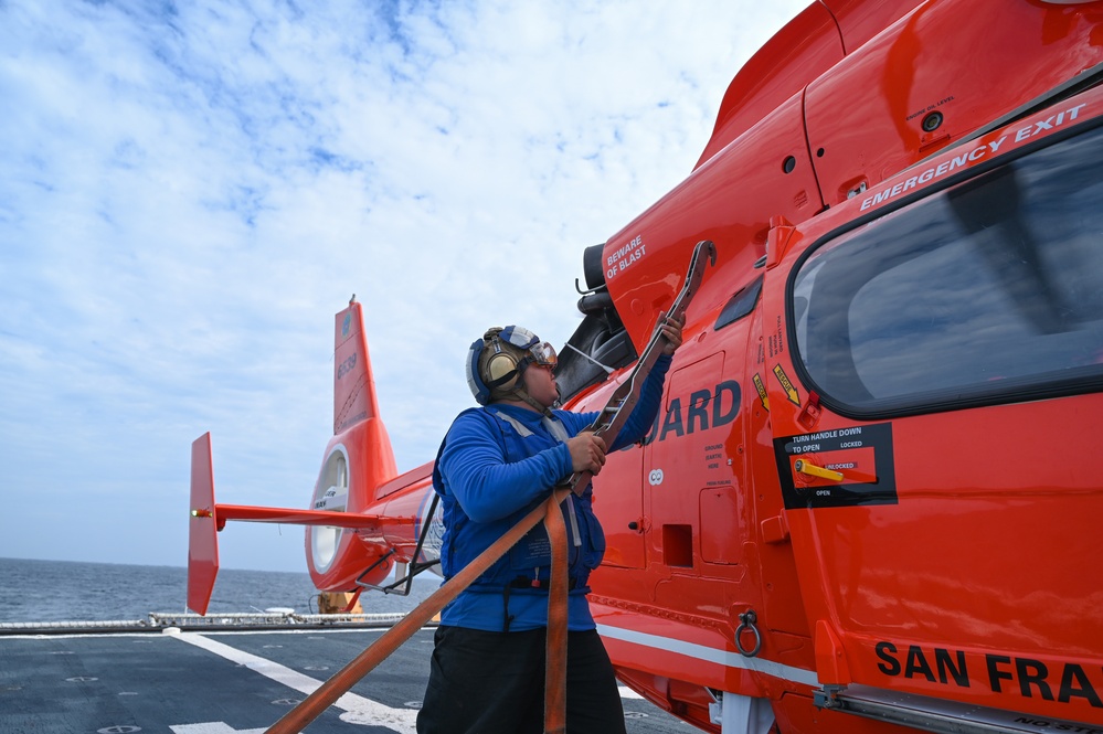 A Coast Guard crew member in a blue flight suit and hearing protection is performing maintenance or pre-flight checks on a bright orange U.S. Coast Guard helicopter on the deck of a ship at sea. The helicopter is partially visible with 'SAN FRA' (San Francisco) and 'GUARD' markings, along with emergency exit and door operation labels. A second orange Coast Guard helicopter is visible in the background on the same flight deck.