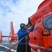 A Coast Guard crew member in a blue flight suit and hearing protection is performing maintenance or pre-flight checks on a bright orange U.S. Coast Guard helicopter on the deck of a ship at sea. The helicopter is partially visible with 'SAN FRA' (San Francisco) and 'GUARD' markings, along with emergency exit and door operation labels. A second orange Coast Guard helicopter is visible in the background on the same flight deck.
