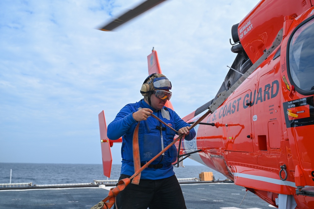 A U.S. Coast Guard crew member in a blue jacket is working on a flight deck, handling tie-down straps or securing lines attached to a bright orange Coast Guard helicopter. The scene takes place on what appears to be the deck of a Coast Guard cutter at sea, with open ocean visible in the background. The helicopter's rotor blades are spinning, indicating the aircraft is either preparing for launch or has recently landed.