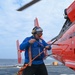 A U.S. Coast Guard crew member in a blue jacket is working on a flight deck, handling tie-down straps or securing lines attached to a bright orange Coast Guard helicopter. The scene takes place on what appears to be the deck of a Coast Guard cutter at sea, with open ocean visible in the background. The helicopter's rotor blades are spinning, indicating the aircraft is either preparing for launch or has recently landed.