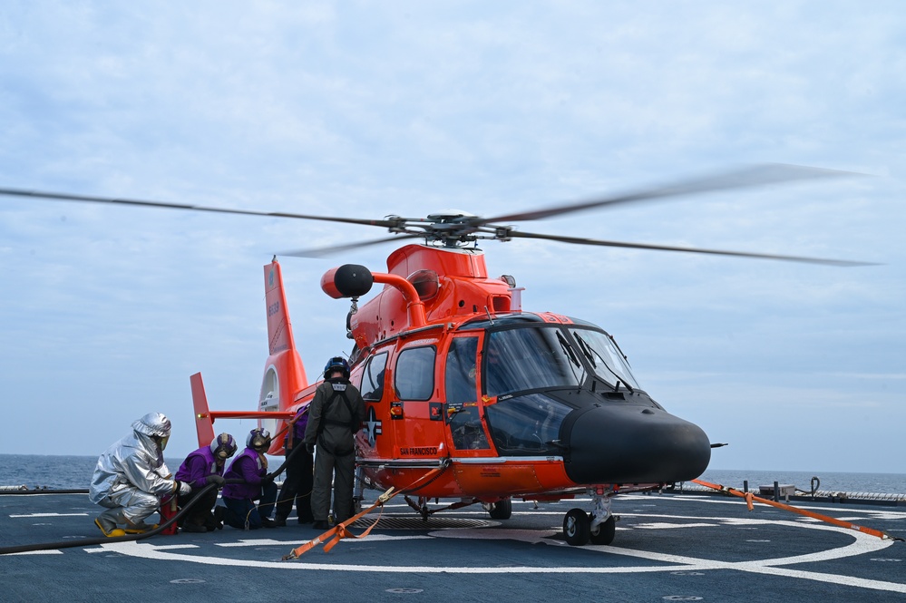 A bright orange U.S. Coast Guard MH-65 Dolphin helicopter sits on a ship's flight deck with its rotors spinning. Several crew members in various protective gear, including silver fire-resistant suits and purple flight deck jerseys, are gathered near the aircraft. The text 'SAN FRANCISCO' is visible on the helicopter's fuselage, indicating its home station.