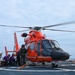 A bright orange U.S. Coast Guard MH-65 Dolphin helicopter sits on a ship's flight deck with its rotors spinning. Several crew members in various protective gear, including silver fire-resistant suits and purple flight deck jerseys, are gathered near the aircraft. The text 'SAN FRANCISCO' is visible on the helicopter's fuselage, indicating its home station.