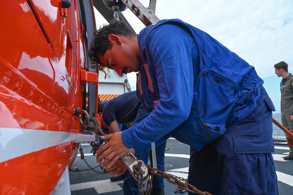 A person in blue coveralls/flight deck uniform is securing a red and white helicopter to a ship's flight deck using chains and tie-down hardware. The scene takes place on what appears to be a Coast Guard or naval vessel's flight deck, with at least two other personnel visible in the background. The red aircraft bears markings consistent with a U.S. Coast Guard helicopter.