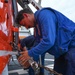 A person in blue coveralls/flight deck uniform is securing a red and white helicopter to a ship's flight deck using chains and tie-down hardware. The scene takes place on what appears to be a Coast Guard or naval vessel's flight deck, with at least two other personnel visible in the background. The red aircraft bears markings consistent with a U.S. Coast Guard helicopter.