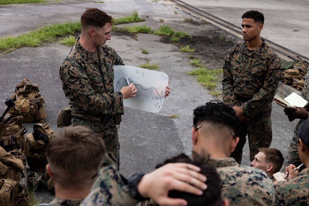 A military briefing is taking place outdoors on a paved surface, where a Marine in MARPAT camouflage uniform is presenting a hand-drawn tactical diagram on a whiteboard to a group of fellow Marines. The presenter appears to be a non-commissioned officer or junior officer conducting an operations order or mission briefing. Several Marines are seated on the ground taking notes while others stand attentively.
