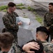 A military briefing is taking place outdoors on a paved surface, where a Marine in MARPAT camouflage uniform is presenting a hand-drawn tactical diagram on a whiteboard to a group of fellow Marines. The presenter appears to be a non-commissioned officer or junior officer conducting an operations order or mission briefing. Several Marines are seated on the ground taking notes while others stand attentively.
