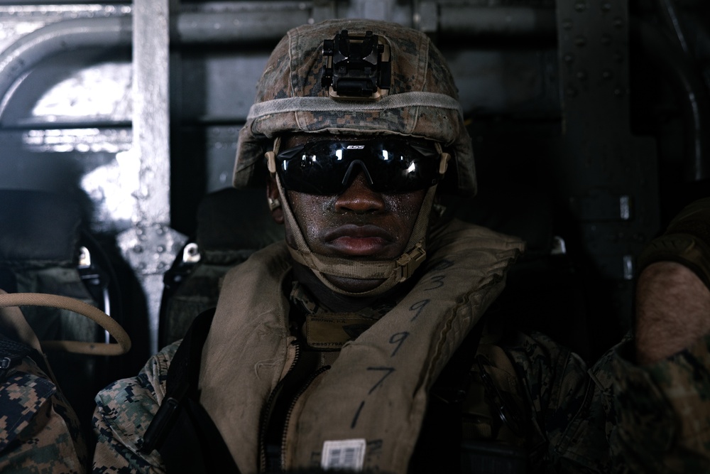 A military service member wearing MARPAT digital camouflage uniform and helmet is seated inside what appears to be a naval vessel or amphibious craft. The individual is equipped with tactical ballistic eyewear (ESS brand), a night vision mount on the helmet, and an inflatable life vest marked with the numbers 3, 9, 7. The image is a close-up portrait-style photograph taken in a dimly lit interior environment.