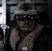 A military service member wearing MARPAT digital camouflage uniform and helmet is seated inside what appears to be a naval vessel or amphibious craft. The individual is equipped with tactical ballistic eyewear (ESS brand), a night vision mount on the helmet, and an inflatable life vest marked with the numbers 3, 9, 7. The image is a close-up portrait-style photograph taken in a dimly lit interior environment.