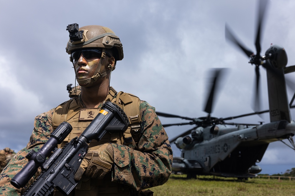 A U.S. Marine in full combat gear stands in the foreground with face camouflage paint, tactical helmet with night vision mount, and an assault rifle with scope. Behind the Marine, a CH-53E Super Stallion helicopter with spinning rotors is visible, marked with 'MARINES' on its fuselage. The image appears to be taken during a field exercise or operational deployment.