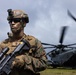 A U.S. Marine in full combat gear stands in the foreground with face camouflage paint, tactical helmet with night vision mount, and an assault rifle with scope. Behind the Marine, a CH-53E Super Stallion helicopter with spinning rotors is visible, marked with 'MARINES' on its fuselage. The image appears to be taken during a field exercise or operational deployment.