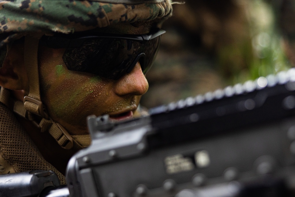 A close-up photograph of a military service member wearing MARPAT (Marine pattern) camouflage gear, tactical eyewear, and face paint in green and brown tones. The subject is positioned behind what appears to be a firearm, suggesting a tactical or training scenario in an outdoor environment. The image has a shallow depth of field, keeping the subject's face in sharp focus while blurring the foreground weapon and background.