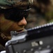 A close-up photograph of a military service member wearing MARPAT (Marine pattern) camouflage gear, tactical eyewear, and face paint in green and brown tones. The subject is positioned behind what appears to be a firearm, suggesting a tactical or training scenario in an outdoor environment. The image has a shallow depth of field, keeping the subject's face in sharp focus while blurring the foreground weapon and background.
