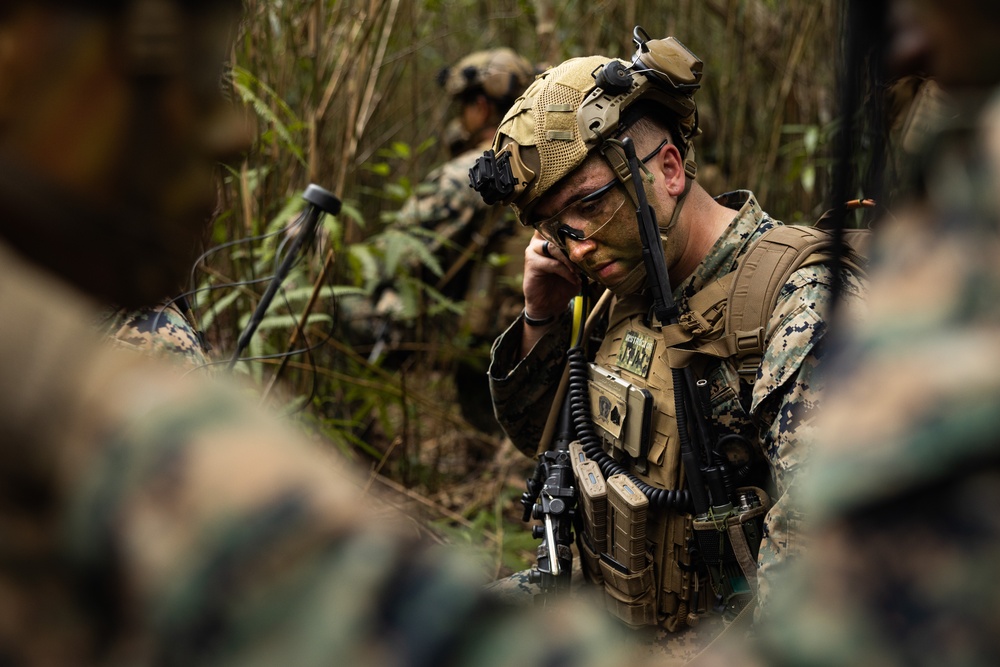 A U.S. Marine in full combat gear is using a radio handset in a dense jungle or forested environment. He is wearing MARPAT digital camouflage, a tan tactical helmet with night vision mount, ballistic glasses, and a heavily loaded plate carrier vest. Additional personnel in similar uniforms are visible in the background.