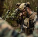 A U.S. Marine in full combat gear is using a radio handset in a dense jungle or forested environment. He is wearing MARPAT digital camouflage, a tan tactical helmet with night vision mount, ballistic glasses, and a heavily loaded plate carrier vest. Additional personnel in similar uniforms are visible in the background.