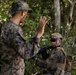 Two military personnel in woodland camouflage uniforms are engaged in what appears to be a tactical field exercise in a forested environment. One soldier wearing a tactical helmet and body armor is kneeling and holding a firearm, while the other is standing and using a hand signal. The setting suggests a training scenario or patrol operation in dense vegetation.