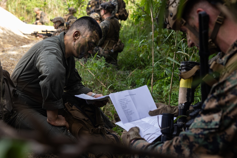 Two military personnel in camouflage uniforms and face paint are reviewing printed documents in a forested outdoor setting. Additional soldiers are visible in the background in tactical positions. The scene appears to depict a military field exercise or training operation.