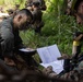 Two military personnel in camouflage uniforms and face paint are reviewing printed documents in a forested outdoor setting. Additional soldiers are visible in the background in tactical positions. The scene appears to depict a military field exercise or training operation.