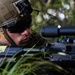 A U.S. Marine in full tactical gear is positioned low in vegetation, aiming down the scope of an assault rifle. The soldier is wearing a tan tactical helmet with night vision device mount, dark ballistic sunglasses, and MARPAT camouflage uniform. A yellow training/safety tag is visible on the weapon, suggesting this may be a training exercise.