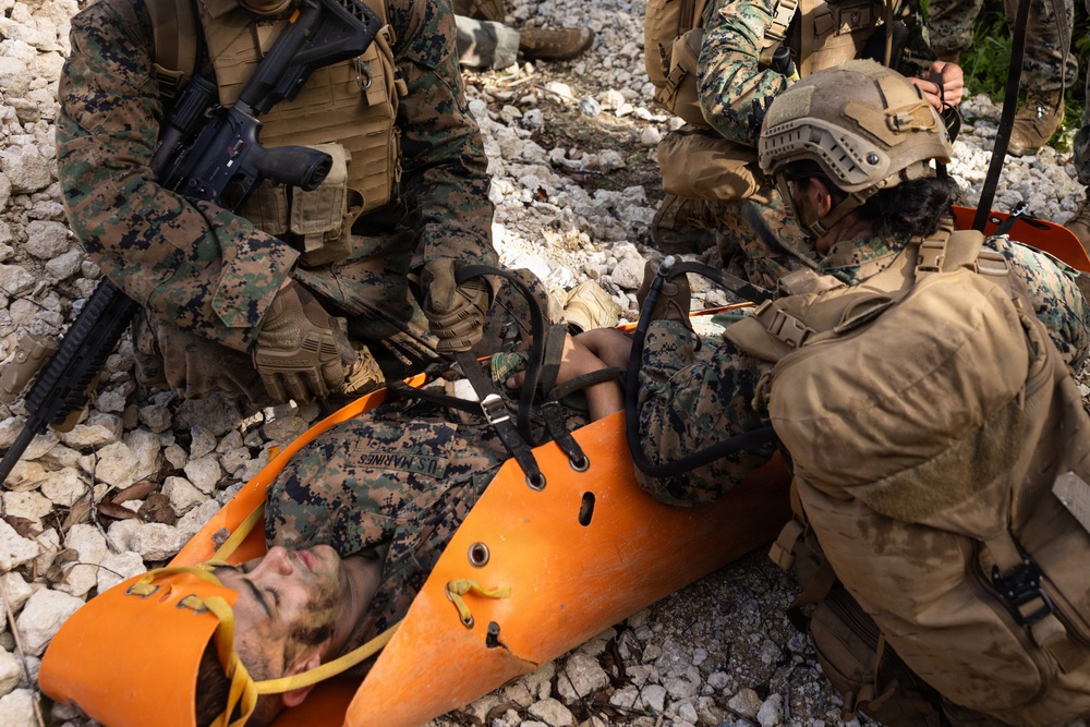 U.S. Marines are conducting a casualty evacuation (CASEVAC) drill or real-world medical response, with one Marine secured to an orange Sked stretcher on rocky terrain. Multiple personnel in MARPAT digital camouflage uniforms and tactical gear are attending to the casualty. The scene appears to be a training exercise or field medical operation in an outdoor rocky environment.