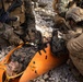 U.S. Marines are conducting a casualty evacuation (CASEVAC) drill or real-world medical response, with one Marine secured to an orange Sked stretcher on rocky terrain. Multiple personnel in MARPAT digital camouflage uniforms and tactical gear are attending to the casualty. The scene appears to be a training exercise or field medical operation in an outdoor rocky environment.