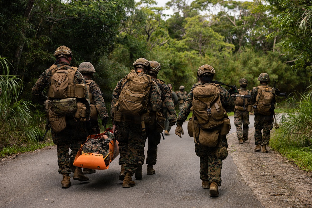 A group of military personnel in full combat gear are moving along a paved road through a densely vegetated area. Two soldiers on the left are carrying a person on an orange rescue/medical litter, suggesting a casualty evacuation exercise or real medical emergency. The scene appears to be a training exercise given the organized movement and mixed equipment visible.
