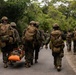 A group of military personnel in full combat gear are moving along a paved road through a densely vegetated area. Two soldiers on the left are carrying a person on an orange rescue/medical litter, suggesting a casualty evacuation exercise or real medical emergency. The scene appears to be a training exercise given the organized movement and mixed equipment visible.
