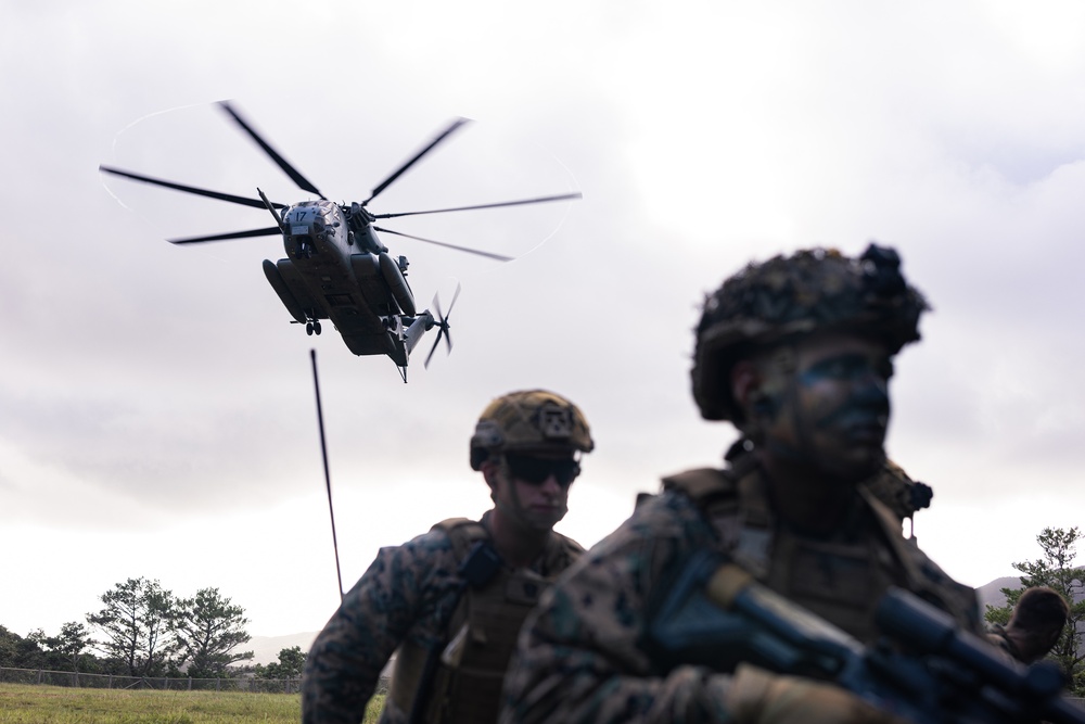 Two military personnel in camouflage uniforms and tactical gear are positioned in the foreground while a large military helicopter hovers in the background. The soldier in the right foreground has camouflage face paint applied, and both wear helmets with tactical attachments. A rope or cable is visibly suspended from the helicopter, suggesting a rappelling or sling-load operation in progress.
