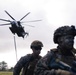 Two military personnel in camouflage uniforms and tactical gear are positioned in the foreground while a large military helicopter hovers in the background. The soldier in the right foreground has camouflage face paint applied, and both wear helmets with tactical attachments. A rope or cable is visibly suspended from the helicopter, suggesting a rappelling or sling-load operation in progress.