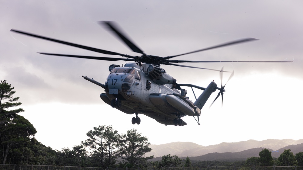 A large military helicopter, identifiable as a CH-53E Super Stallion marked with the number '17', is captured in low hover or landing approach over a fenced area. The aircraft displays motion blur on its main rotor blades and tail rotor, indicating active flight. The background features mountainous terrain, dense tree line, and an overcast sky.