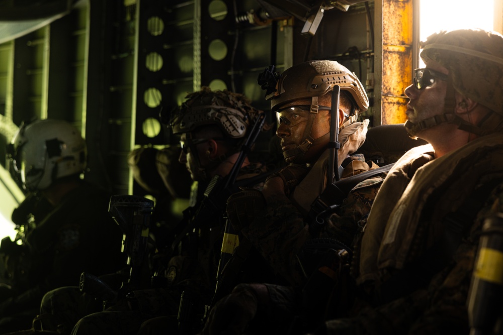 A group of heavily equipped military personnel are seated inside what appears to be a military transport aircraft or helicopter, illuminated by a warm golden light source from the right side. The soldiers are wearing full tactical gear including helmets, body armor, and carrying weapons, suggesting they are preparing for or returning from an operation. The dramatic lighting and tight framing create an intense, cinematic atmosphere.