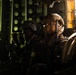 A group of heavily equipped military personnel are seated inside what appears to be a military transport aircraft or helicopter, illuminated by a warm golden light source from the right side. The soldiers are wearing full tactical gear including helmets, body armor, and carrying weapons, suggesting they are preparing for or returning from an operation. The dramatic lighting and tight framing create an intense, cinematic atmosphere.