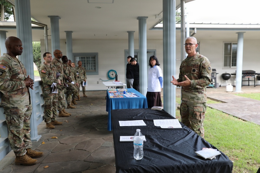 A senior U.S. Army officer in OCP (Operational Camouflage Pattern) uniform addresses a group of soldiers lined up under a covered outdoor patio area. Two information or resource tables are set up with materials, covered in black and blue tablecloths, suggesting a briefing, outreach, or resource fair event. Two civilians are visible in the background near the tables.