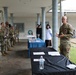A senior U.S. Army officer in OCP (Operational Camouflage Pattern) uniform addresses a group of soldiers lined up under a covered outdoor patio area. Two information or resource tables are set up with materials, covered in black and blue tablecloths, suggesting a briefing, outreach, or resource fair event. Two civilians are visible in the background near the tables.