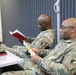 Two U.S. Army soldiers in MultiCam/OCP camouflage uniforms are seated in what appears to be a classroom or briefing room, engaged in reading materials. The soldier in the foreground wears glasses and is reading a small pamphlet or booklet, while the soldier in the background reads a red-covered book. Both wear unit patches on their left shoulders consistent with U.S. Army insignia.