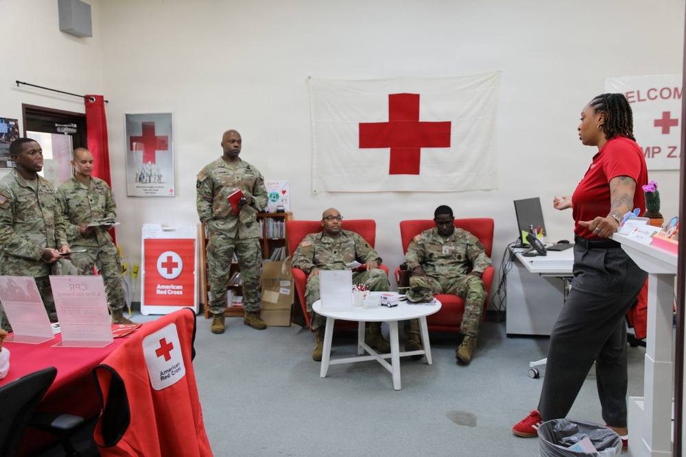 A female American Red Cross representative in a red shirt is presenting or briefing a group of military personnel in camouflage uniforms inside a Red Cross office or service center. The setting appears to be a military installation, likely overseas, based on the partial 'Welcome Camp Za...' signage visible in the background. The room is decorated with Red Cross branding, flags, and informational materials.
