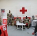 A female American Red Cross representative in a red shirt is presenting or briefing a group of military personnel in camouflage uniforms inside a Red Cross office or service center. The setting appears to be a military installation, likely overseas, based on the partial 'Welcome Camp Za...' signage visible in the background. The room is decorated with Red Cross branding, flags, and informational materials.