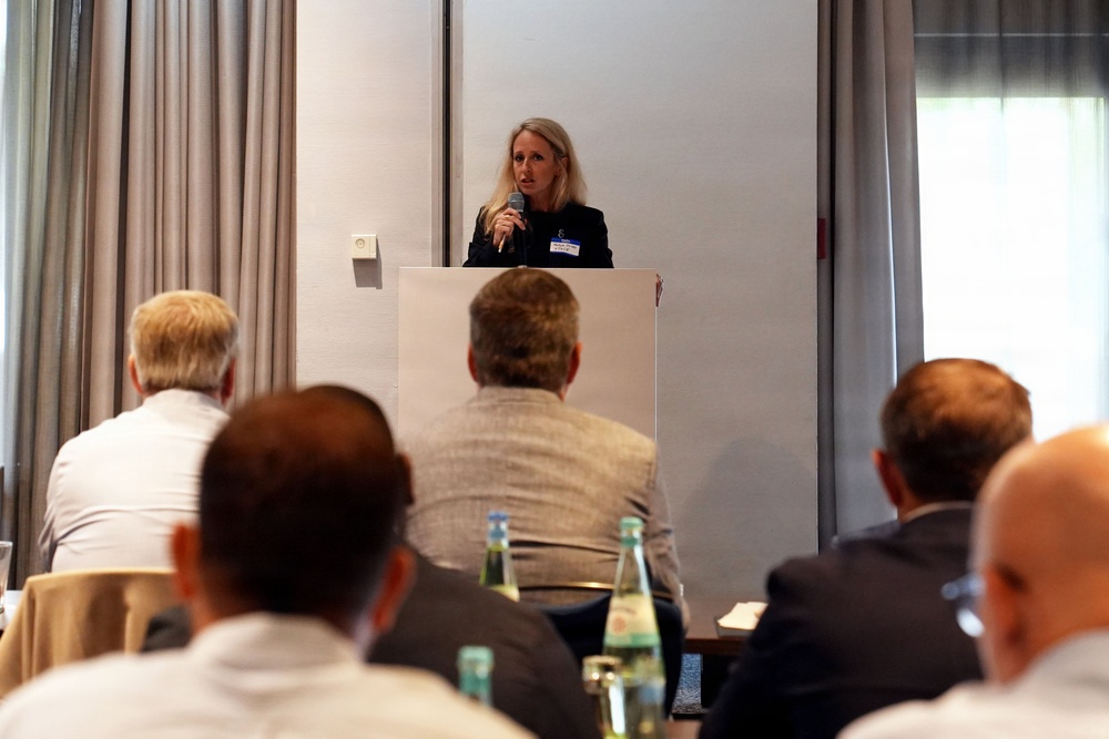A woman with blonde hair is speaking at a podium during what appears to be a professional conference or business meeting. She is holding a microphone and wearing a name badge, addressing an audience of predominantly male attendees seated at tables. The setting appears to be a hotel conference room with curtained windows and neutral decor.